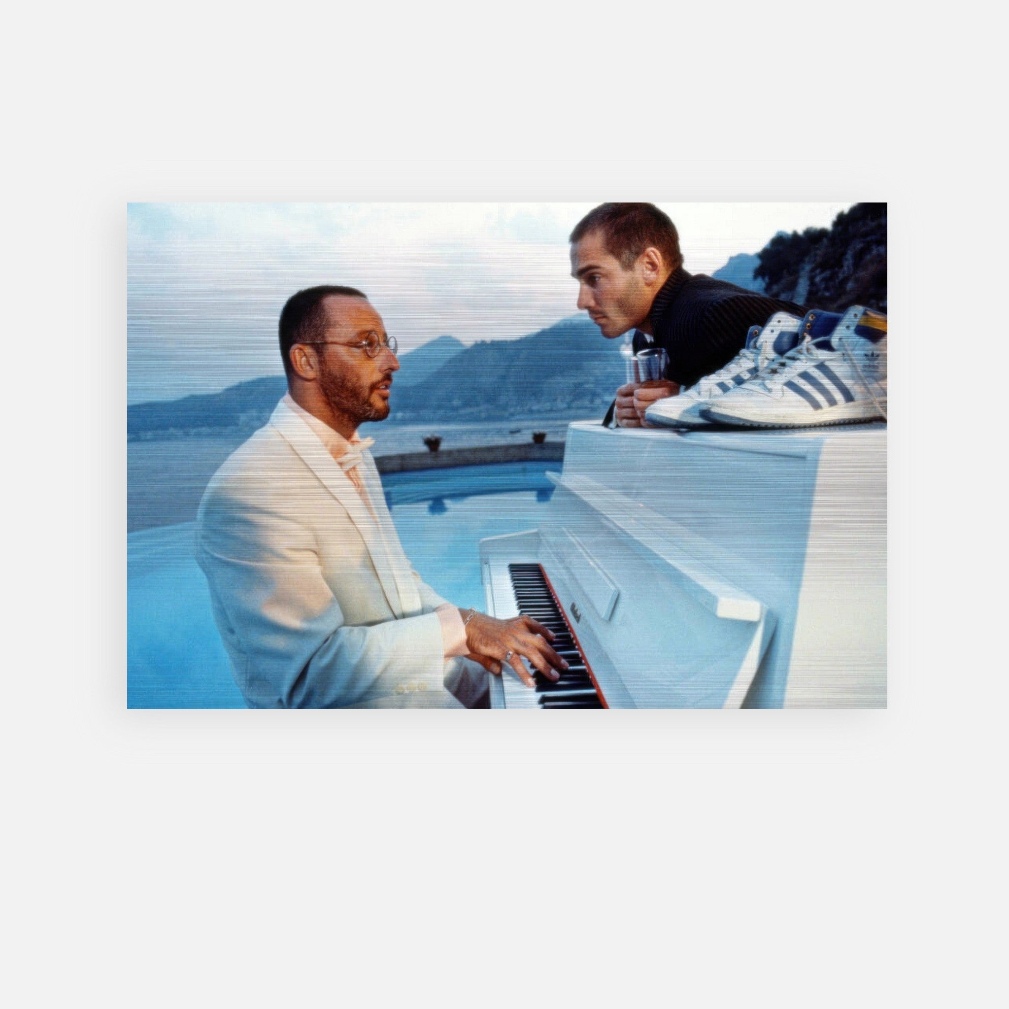 A man in a white suit plays a white piano while another man leans in, with mountains and sneakers in the background.
