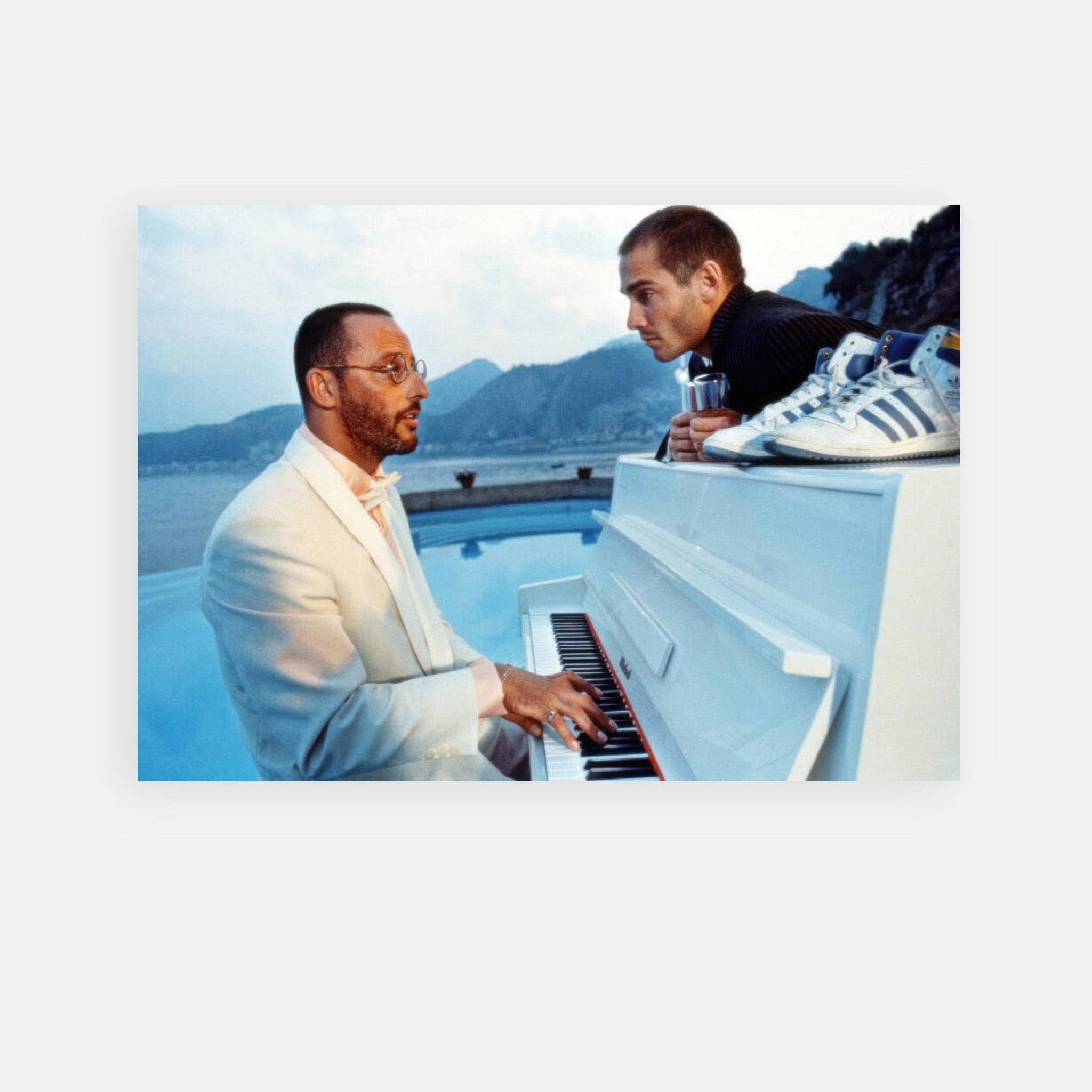 Jean Reno playing a white piano on a scenic coastal balcony with a friend, capturing a moment of artistic charm.