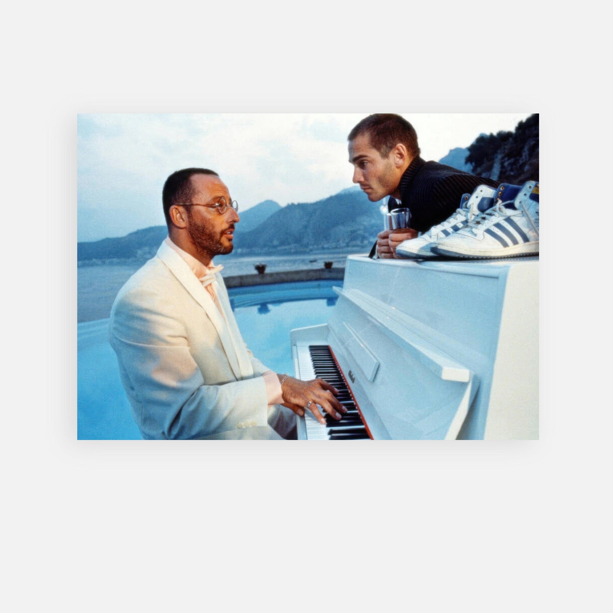 Jean Reno playing a white piano by the coastal sea, with a companion watching, capturing a serene musical moment.