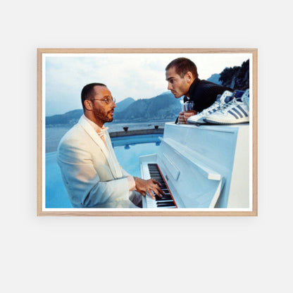 Vintage framed print of Jean Reno playing piano on the set of "The Big Blue" with scenic seaside background.